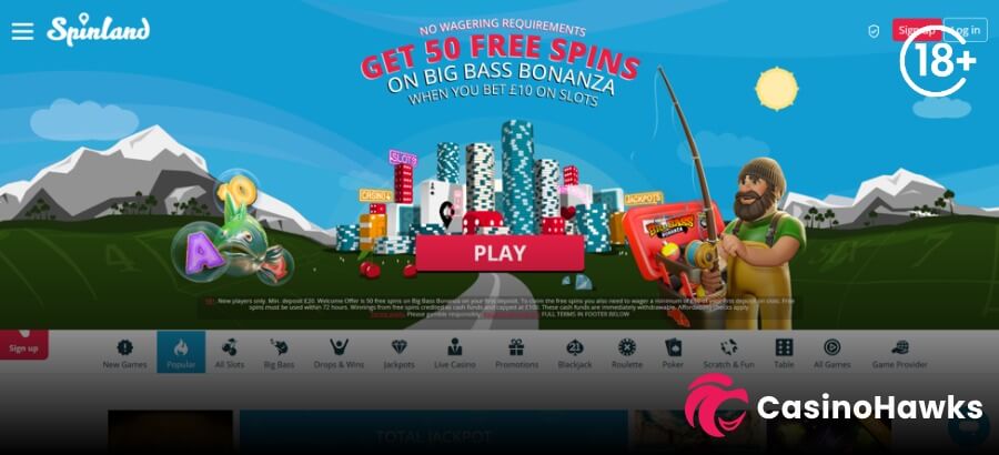 A desktop landing page hero banner for Spinland Casino featuring a illustrated cityscape made of playing cards, slot machines, and chip stacks beneath a bright blue sky with snowy mountains. Bold pink and white headline text reads "NO WAGERING REQUIREMENTS - GET 50 FREE SPINS ON BIG BASS BONANZA WHEN YOU BET £10 ON SLOTS" above a prominent pink "PLAY" button. On the right, Pragmatic Play's fisherman character stands with a fishing rod next to a Big Bass tackle box. An 18+ age restriction badge rests in the top right corner, while fine-print T&C text sits at the bottom of the banner. The lower navigation bar displays filter icons for New Games, Popular, All Slots, Big Bass, Drops & Wins, Jackpots, Live Casino, Blackjack, Roulette, and Game Provider, with the Casino Hawks pink lion co-branding logo in the bottom right corner.