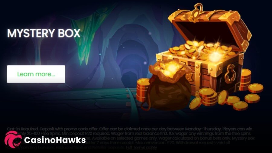 An online casino promotional banner advertising a "MYSTERY BOX" daily bonus offer set inside a dark mine shaft cave with stalactites and train tracks. On the right sits a large, glowing wooden treasure chest overflowing with gold coins, flanked by stacks of gold coins and a brown sack filled with treasure. A white "Learn more..." button rests on the left, and a "CasinoHawks" affiliate logo is watermarked in the bottom left corner over detailed promotional terms and conditions.